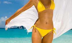Picture of a woman, happy with her abdomen and waist  liposuction procedure she had at Top Plastic Surgeons in beautiful San Jose, Costa Rica.  The woman is wearing a two piece yellow bathing suit and walking through the surf at the beach.